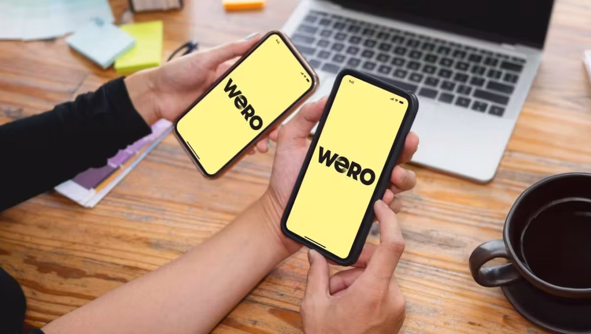 two people holding phones with the word WERO on the center of their screens. there is a wooden table in the background with a laptop and coffee on it.