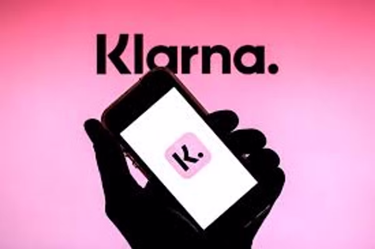 A silhouetted hand holding a phone showing the Klarna app icon, against a pink background with the Klarna wordmark above.