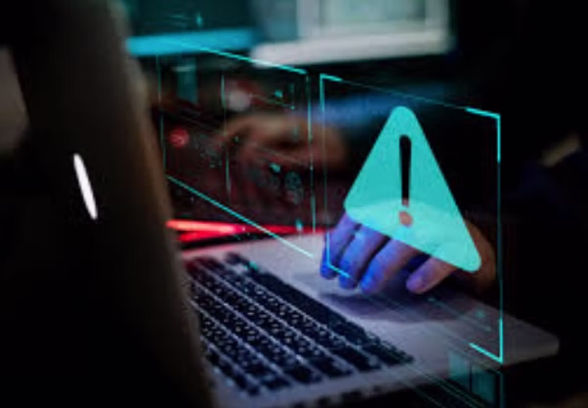 Someone typing at a laptop in a dark room, with a large glowing warning triangle projected above the keyboard.